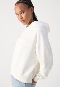 Kreemjas puuvillane kapuutsiga sviiter, millel on vaba lõige, esipaneelil heleroosa logo, nööriga kapuuts ning soonikuga varrukad ja alläär.