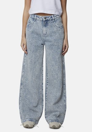 Femme portant un jean bleu à jambes larges et taille haute avec un motif quadrillé texturé et des baskets blanches, debout les bras détendus.