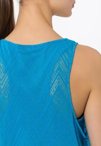 Kvinnas övre rygg och nacke, iförd en ärmlös blå topp med ett chevronmönster i genomskinlig tyg.