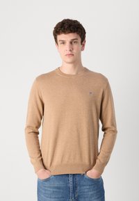 Beige kötött pulóver kerek nyakkal, hosszú ujjal, bordázott mandzsettával és szegéllyel. Kis logó részlet a bal mellkason. Kék farmerral viselve.
