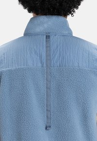 Svetlomodrá flísová bunda s textúrovanou hornou časťou a hladkou dolnou časťou, s nastavovacím pútkom na zadnej strane.