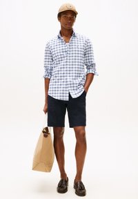 Chemise blanche à carreaux bleus et verts, short marine, mocassins marron et sac beige avec un logo, associés à une casquette beige.