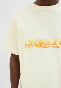 Korte mouwen, licht crème t-shirt van zachte katoen, met een getextureerde oranje gradient-print en de tekst "project TM CREATIVE DEPT."