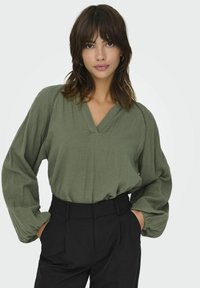 Olivgrüne Bluse mit V-Ausschnitt, langen Ärmeln und strukturiertem Stoff, kombiniert mit hoch taillierten schwarzen Hosen.