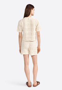 Conjunto de dos piezas de ganchillo en color beige cremoso, que incluye una blusa corta de manga corta y un pantalón corto de tiro alto con bordes ondulados.