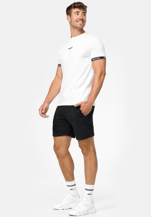 Hvid T-shirt med sorte ribbede ærmer, der har "INDICODE" logoet, parret med sorte shorts og hvide kondisko med stribede sokker.