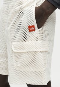 Shorts cargo en maille blanche avec poche latérale et écusson du logo rouge The North Face, portés avec une main dans la poche.