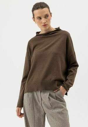 Woman wearing a brown loose sweater and gray pleated trousers, standing with one hand in pocket, looking to the side against a light background.