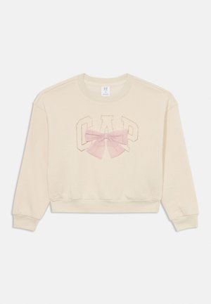 Sweatshirt beige avec un design court, arborant le logo "GAP" brodé et un nœud en tulle rose au centre. Tissu doux et texturé.