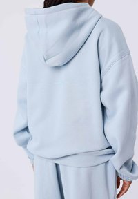 Persona che indossa una felpa leggera blu e pantaloni della tuta abbinati, vista da dietro con le mani rilassate ai lati.