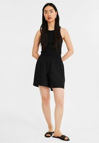 Débardeur noir à côtes associé à un short noir à taille élastique. Le short a une coupe ample et une texture lisse. Porté avec des sandales à glissière noires.