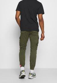 Khaki cargo pants met zijzakken, slankere pasvorm, elastische boorden en een rode label. Draagt een zwart t-shirt en grijze sportchoenen.