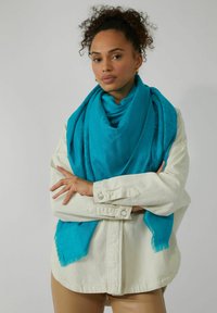 Écharpe turquoise en tissu doux, avec un motif subtil et des bords effilochés, enroulée autour des épaules sur une veste crème.