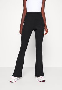 Černé rozšířené kalhoty z elastického materiálu s vysokým pasem a rozparkem na nohavicích, v kombinaci s bílými teniskami s barevnými akcenty.