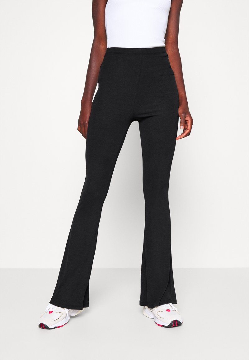 Černé rozšířené kalhoty z elastického materiálu s vysokým pasem a rozparkem na nohavicích, v kombinaci s bílými teniskami s barevnými akcenty.