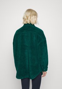 Tealfärgad fleecejacka med en mjuk, texturerad yta, hög krage och avkopplad passform, med rundad nederkant och detaljer med knappar vid ärmarna.