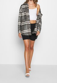 Overmaat zwart-wit geruit shirtjack met knopen, gecombineerd met een witte crop top en zwarte shorts. Strappy witte hakken.