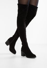 Stivali neri sopra il ginocchio realizzati in morbida suede, con tacco spesso e design aderente che abbraccia la gamba. Cerniera laterale visibile.