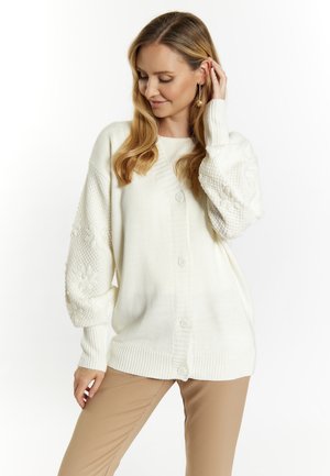 Cardigan bianco lavorato a maglia con maniche lunghe e testurizzate e colletto rotondo. Presenta una chiusura con bottoni e orlo a coste. Indossato su pantaloni beige.