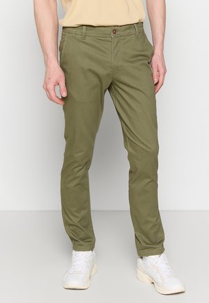 Hombre vestido con pantalones ajustados verde oliva y zapatillas blancas, de pie contra un fondo claro y liso.