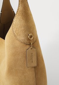 Suede handbag in light brown with a rounded shape, featuring a stitched edge and a gold hardware tag. Notable soft texture and simple design.