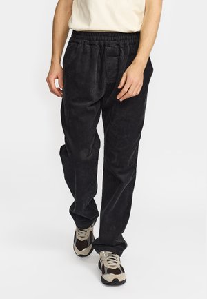 Uomo con pantaloni in velluto a coste neri e sneaker beige, marroni e nere, in piedi davanti a uno sfondo bianco uniforme.