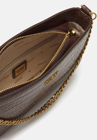 Brun faux læder taske med krokodillemønster, guld kædetraspe, lynlås lukning og beige interiør med "GUESS" logo.