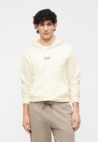 Jasnożółta bluza z kapturem z przednią kieszenią kangurka, ściągaczem na kapturze i detalem z logo. Połączona z beżowymi dresami z regulowanym sznurkiem w pasie.