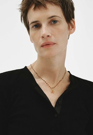 Mujer con cabello corto y castaño, vistiendo una blusa negra y un collar de cuentas con un colgante de letra "R" en dorado sobre un fondo blanco.