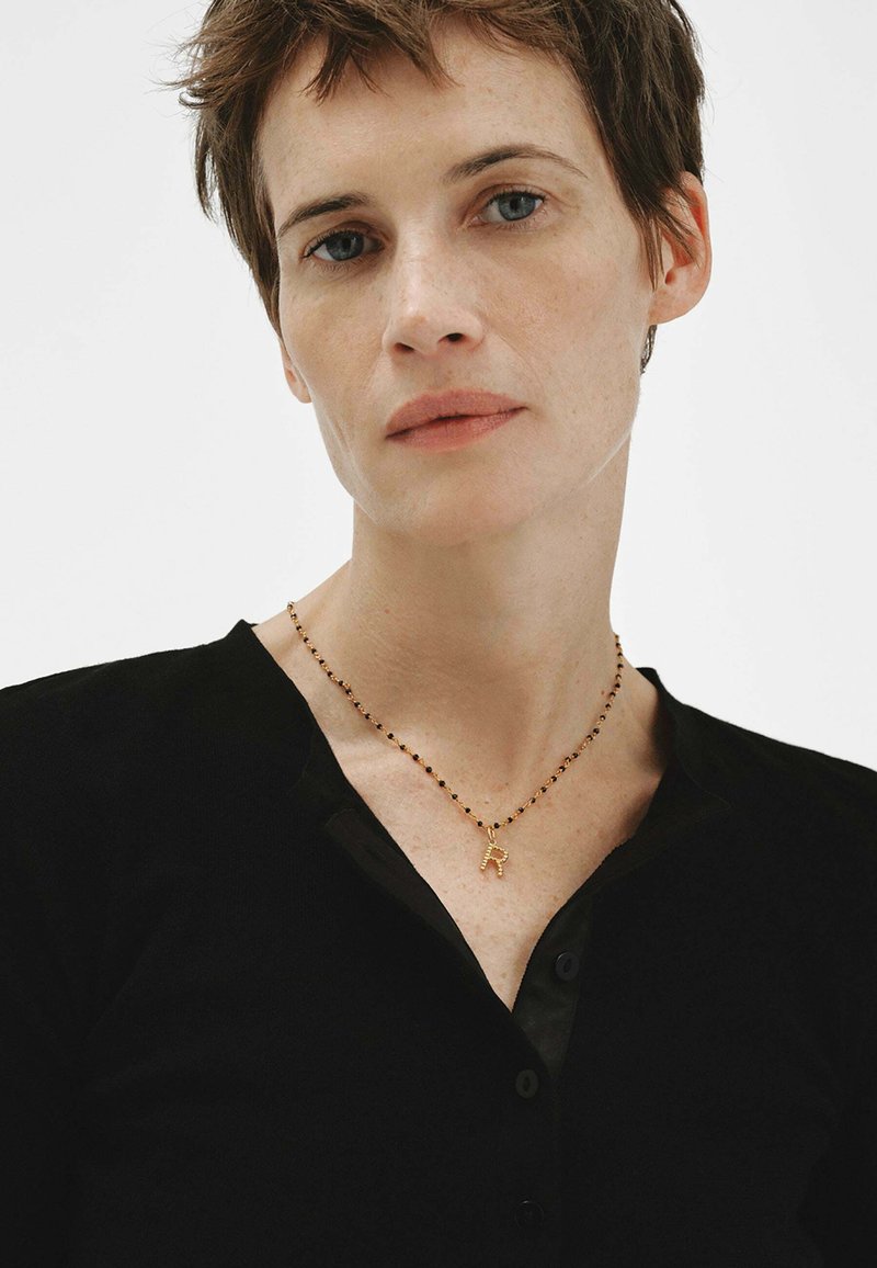 Mujer con cabello corto y castaño, vistiendo una blusa negra y un collar de cuentas con un colgante de letra "R" en dorado sobre un fondo blanco.