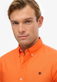 Pomarańczowa koszula z guzikami wykonana z bawełny. Posiada kołnierzyk, listwę guzikową oraz haftowane logo po lewej stronie klatki piersiowej. Gładka tekstura.