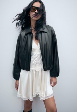 Schwarze Lederbomberjacke mit einem Reißverschluss, getragen über einem weißen Kleid mit Spitzenbesatz. Das Model posiert vor einer neutralen Wand.