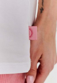 Bílá bavlněná tričko s kulatým lemem; má růžovou textilní etiketu se slunečním designem na boku. Detailní záběr ruky držící tričko.