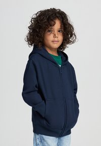 Felpa con zip di colore blu navy, realizzata in tessuto morbido, con tasca a marsupio e polsini a coste. Indossata sopra una maglietta verde, con un taglio rilassato.