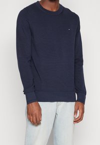 Pull tricoté manches longues bleu foncé avec col rond et petit logo rouge, blanc et bleu sur la poitrine gauche, porté avec un jean bleu clair.