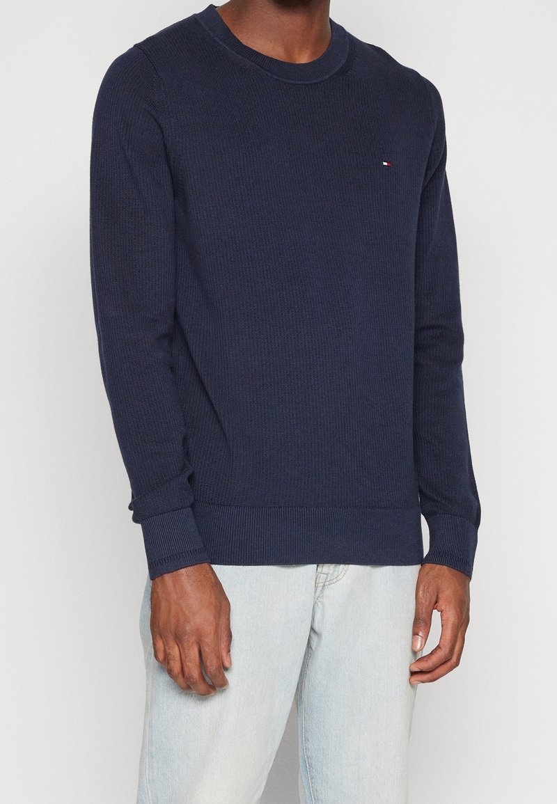 Pull tricoté manches longues bleu foncé avec col rond et petit logo rouge, blanc et bleu sur la poitrine gauche, porté avec un jean bleu clair.