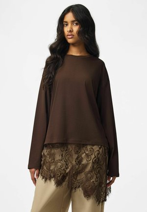 Femme aux longs cheveux foncés portant un haut marron ample à manches longues superposé à une jupe beige avec des détails en dentelle foncée à l'ourlet.