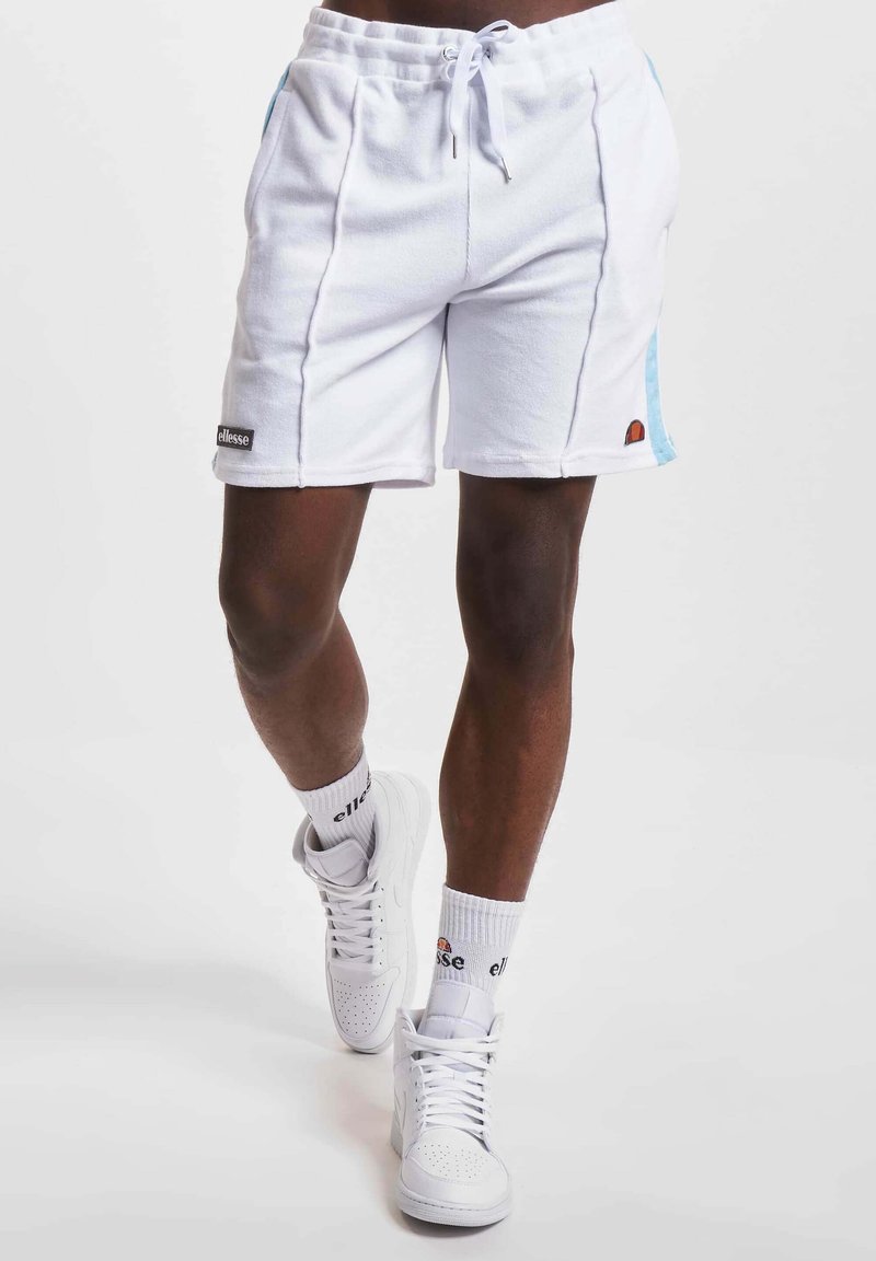 Homme portant un short de sport blanc avec des rayures latérales bleues, des baskets montantes blanches et des chaussettes blanches avec des logos de marque marchant en avant.