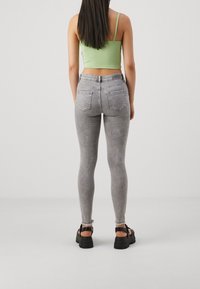 Femme aux longs cheveux foncés portant un crop top vert, un jean skinny gris et des sandales à plateforme noires, de dos.