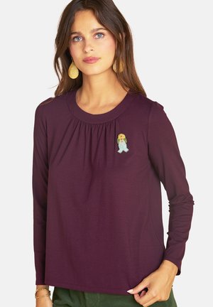 Woman with long brown hair wears a purple long-sleeve top with a small embroidered flower patch and gold textured earrings.