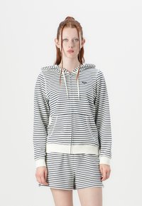 Veste à capuche zippée rayée en bleu marine et blanc avec un cordon de serrage, poches avant et poignets côtelés. Associée à un short assorti.