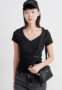 Camiseta negra de cuello en V combinada con pantalones vaqueros grises y un pequeño bolso cruzado negro; el bolso tiene un cierre de cremallera y un acento de logo. Se lleva una gorra negra.