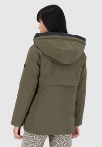 Giacca isolante verde oliva con cappuccio nero, caratterizzata da una vestibilità comoda, cuciture piatte e un'apertura sul retro. Pantaloni a fantasia visibili sotto.