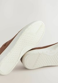 Bruine schoeisel met een gestructureerde bovenkant en witte rubberen zolen met een geometrisch profiel. De merknaam "NEXT" is zichtbaar op de zool.