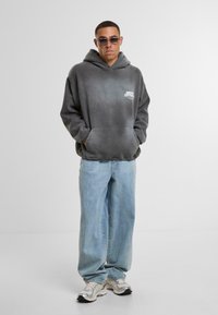 Szary hoodie tie-dye z kieszenią z przodu i logo na lewej piersi, w połączeniu z jasnoniebieskimi luźnymi dżinsami i szarymi trampkami.