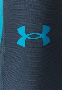 Logo bleu Under Armour sur un tissu gris foncé texturé avec des coutures visibles le long du bord gauche.