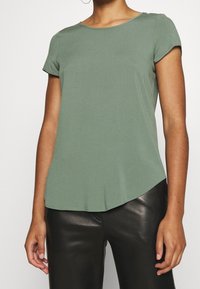 Krátký rukáv, top tlumené zelené barvy, kulatý výstřih, měkký materiál, pohodlný střih, zakřivený lem, spárováno s černými slim-fit kalhotami.