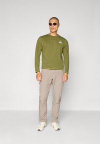 Olivgrünes Langarmhemd mit einem dreieckigen Logo, beiges Hosen und helle Schuhe. Model trägt beigen Sonnenbrillen. Einfaches Design, funktioneller Stil.