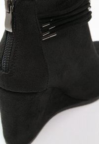 Botín negro de ante con cuña y cierre de cremallera, adornado con cordones decorativos. Textura suave con forma redondeada.