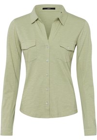 Camicia a maniche lunghe verde chiaro realizzata in materiale morbido. Presenta un colletto, due tasche frontali e chiusura con bottone, con una texture sottile.